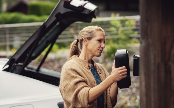 Blond dame med beige ullgenser plasserer sort elbillader på brun husvegg, med hvit bil i bakgrunnen.